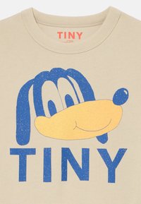 Beige T-Shirt mit einem blauen Cartoon-Hundegesicht und dem Wort "TINY" in fetten blauen Buchstaben. Der Stoff hat eine glatte Textur und einen Rundhalsausschnitt.