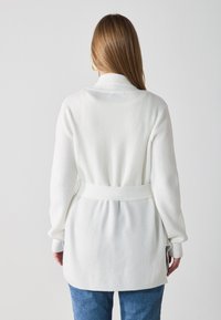 White ribbed knit cardigan with a waist tie, long sleeves, and a shawl collar. Soft texture and casual fit, featuring side slits.
