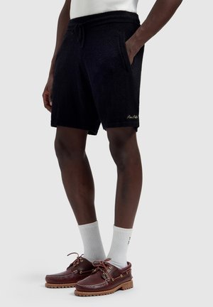 Male model wearing black textured shorts with pockets, white crew socks, and brown leather boat shoes, standing against a plain background.