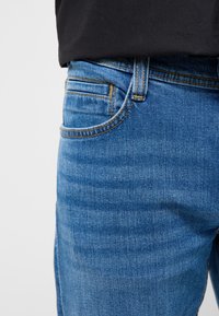 Detail modrých riflí v oblasti vrecka a pásu so žltým prešívaním, nosené s čiernou košeľou zastrčenou dovnútra.