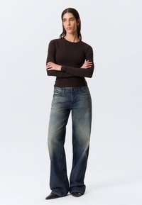 Brūns krekls ar gariem piedurknēm, kopā ar izbalējušām zilām platām džinsa biksēm. Apģērbs ir vaļīga piegriezuma ar gludiem, teksturētiem audumiem.