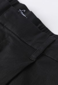 Black fabric waistband of a pair of pants, featuring smooth texture, gathered elastic detail, and a label inside.