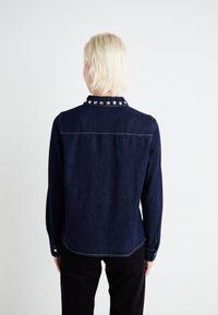 Tume sinine denim-särk nööbitava kraega, millel on hõbedased nupukesed, pikad varrukad ja kõver hem, demonstreerides klassikalist ja struktureeritud disaini.