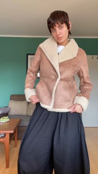 Giacca in suede marrone con un ampio colletto avorio, dotata di chiusura con un singolo bottone e dettagli avorio sui polsini. Il modello indossa pantaloni neri larghi.