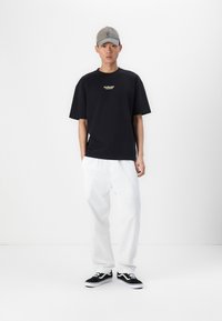 Tricou din bumbac negru cu logo-ul auriu "Hollister", pantaloni albi lejeri și tenisi negri Vans. Modelul poartă o șapcă gri.