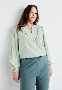 Light green long-sleeve blouse with a V-neck, pleated front, and decorative stitching. Paired with teal fitted pants featuring a drawstring.