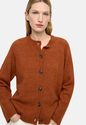 Kobieta o blond włosach, ubrana w rdzawobrązowy sweter rozpinany na guziki, z rękami w kieszeniach białych spodni.