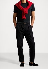 Pantaloni neri, t-shirt nera aderente, maglione rosso in maglia appoggiato sulle spalle, scarpe nere senza lacci, texture liscia e design casual.