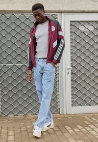 Casaco corta-vento bordeaux com detalhes em preto e branco, suéter canelado cinza claro, calças de ganga largas azul claro e ténis com um design transparente.