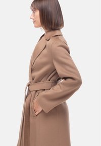Cappotto avvolgente in misto lana di colore marrone con cintura, dotato di colletto a scialle, maniche lunghe e tasche laterali. Tessuto liscio senza fantasie.