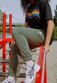 Persoon in groene Nike joggingbroeken en een shirt met regenboogafbeelding klimt op een rode speelstructuur, grijpt een touw, en draagt witte sneakers en gestreepte sokken.