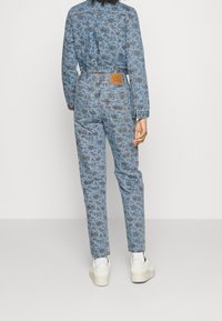 Denim jumpsuit med blomsterdesign i blå og pink. Har en tætsiddende talje, lange ærmer og baglommer. Hvide sneakers fuldender looket.