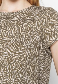 Haut vert avec un motif de feuilles blanches. Col rond, manches courtes, texture lisse et silhouette ajustée.