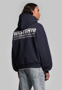 Hoodie oversize bleu marine en tissu doux, avec un grand logo blanc imprimé au dos. Accompagné d'un jean en denim clair.