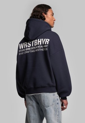 Felpa oversize blu navy realizzata in tessuto morbido, con un grande logo bianco stampato sulla schiena. Accompagnata da jeans in denim chiaro.