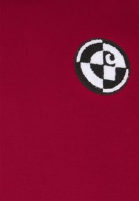Red shirt fabric with a smooth texture features a circular black and white embroidered logo on the front, with geometric patterns.