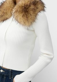 Vit ribbade zip-up tröja med en fluffig brun fuskpälskrage. Långa ärmar och figursydd design med synliga sömdetaljer.