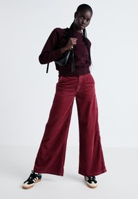 Pantaloni largi din corduroy de culoare maronie, cu un finisaj texturat, asortați cu un hanorac burgund și adidași negri, completând ținuta cu o geantă elegantă.