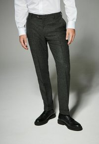 Dunkelgraue, maßgeschneiderte Hose aus strukturiertem Stoff, mit schlanker Passform und subtil gemustertem Fleck, kombiniert mit schwarzen Schuhen.
