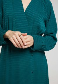 Robe en teinte bleu canard avec un motif texturé, des poignets boutonnés et un décolleté en V profond. Les mains sont croisées, mettant en valeur une bague en or à la main gauche.