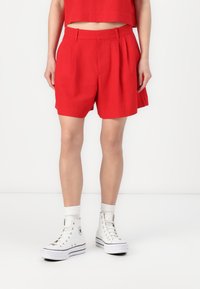 Shorts plissés rouges en tissu léger avec une taille haute. Portés avec des baskets blanches montantes et des chaussettes blanches. Fond uni.