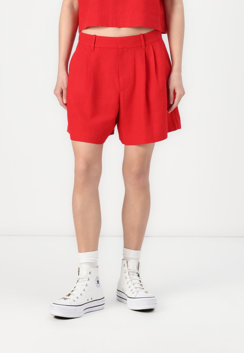 Shorts plissés rouges en tissu léger avec une taille haute. Portés avec des baskets blanches montantes et des chaussettes blanches. Fond uni.