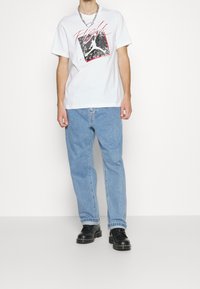Camiseta de algodón blanca con un diseño gráfico en negro y rojo. Llevada con jeans de denim azul claro y zapatos negros con cordones.