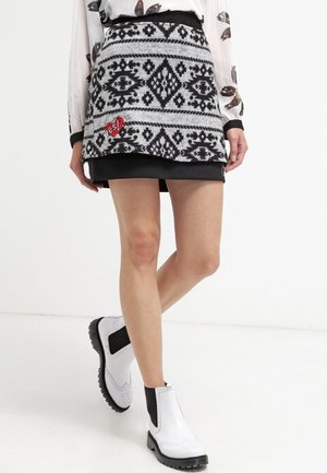 Osoba ubrana w spódnicę w geometryczne czarno-białe wzory z naszywką w kształcie czerwonego serca, przezroczystą białą bluzkę z nadrukiem liści oraz białe botki za kostkę.