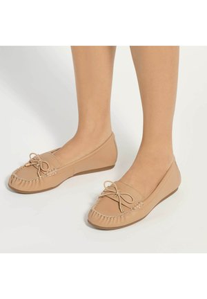 Pieds portant des mocassins en cuir beige avec un nœud devant et des coutures, posés sur une surface blanche.