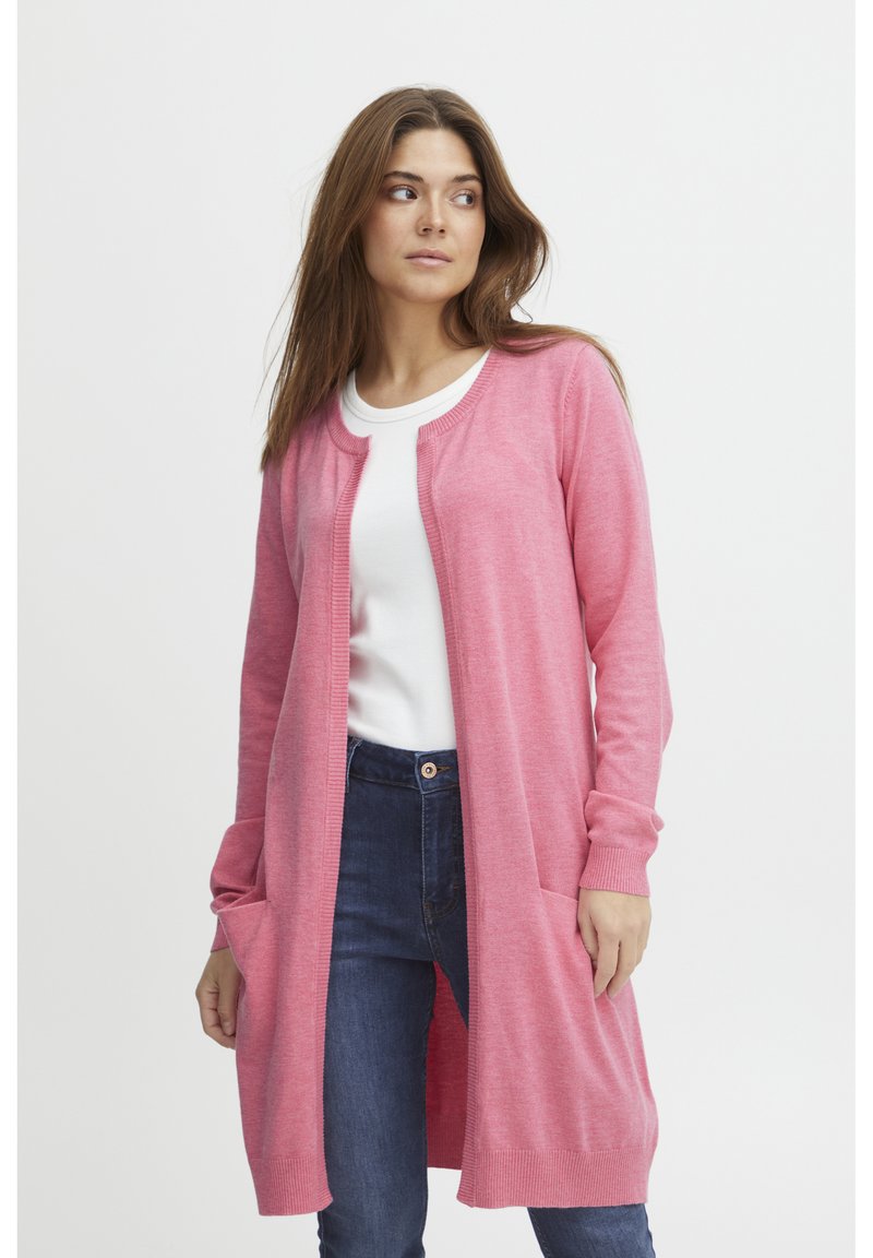 Lichtroze, textuurgebreide cardigan met een ronde halslijn, open voorkant en voorzakken, gedragen over een witte blouse en een blauwe spijkerbroek.