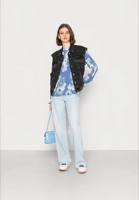 Colete de ganga preto sobre uma camisa azul com estampado floral de mangas compridas, jeans azul claro e ténis brancos com solas rosas. Pequena carteira azul.