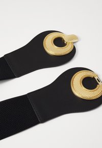 Cinto elástico preto com decoração em hardware circular dourado, apresentando um acabamento texturizado e acentos lisos em cada extremidade.