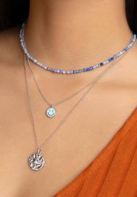 Collane a tre strati: una catena d'argento, un pendente con un sorriso blu e una collana choker di perline multicolori con texture lucide e dimensioni varie.