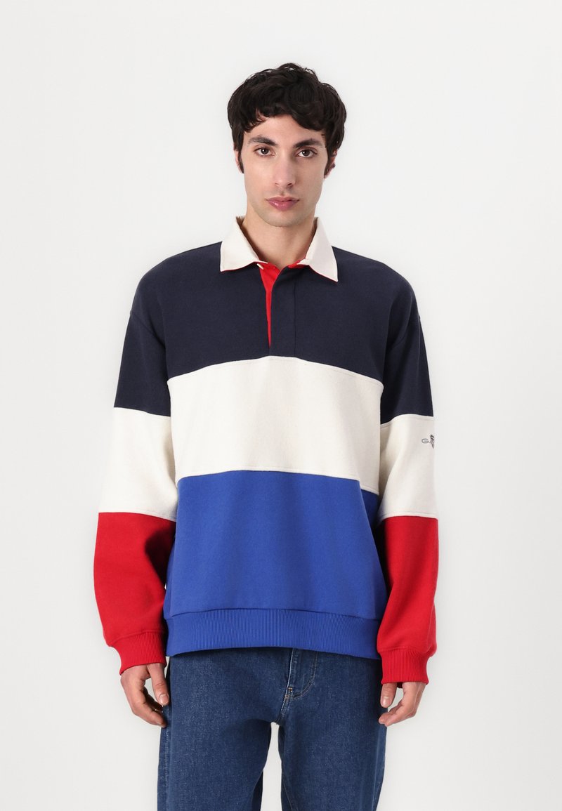 Pullover i marineblå, creme, rød og blå sektioner; har en krave med tre knapper, lange ærmer og ribbede manchetter og kant. Casual design.