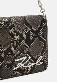 Mustavalkoinen käärmeenahkakuosinen käsilaukku, jossa on sileä pinta ja hopeaketjusangalla varustettu olkahihna. Etuosassa näkyy selkeä valkoinen logo.