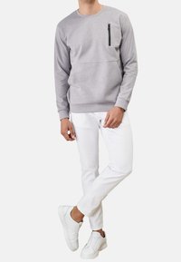 Uomo che indossa una felpa a maniche lunghe grigio chiaro con una tasca con zip nera, pantaloni bianchi e sneakers bianche, in piedi con le gambe incrociate.