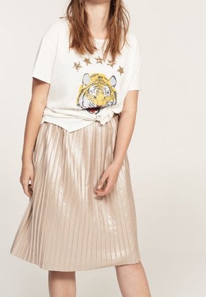 Mujer con camiseta blanca con gráfico de un tigre y estrellas, anudada en la cintura, combinada con una falda midi plisada metálica brillante.