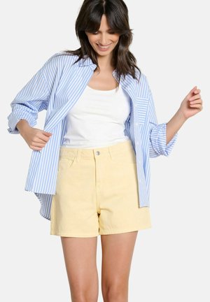 Mujer ajustándose una camisa de rayas azules y blancas sobre una camiseta sin mangas blanca, con pantalones cortos amarillo claro, sonriendo y mirando hacia abajo.