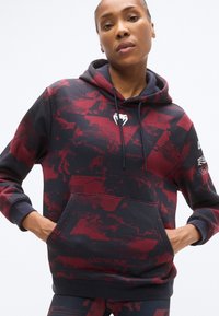 Sweat à capuche à motifs rouge et bleu marine avec poche avant, capuche à cordon et logo sur la poitrine ; fabriqué en tissu doux et texturé.