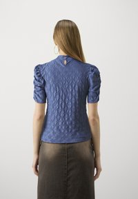 Modrá texturovaná blůza s nabíranými rukávy, zapínáním na knoflík vzadu a výřezem ve tvaru klíčové dírky na krku. Spárována s hnědou sukní.