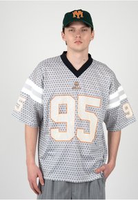 Joven con una camiseta deportiva gris con estampado y el número 95, y una gorra negra con el logo naranja "M" sobre fondo blanco.