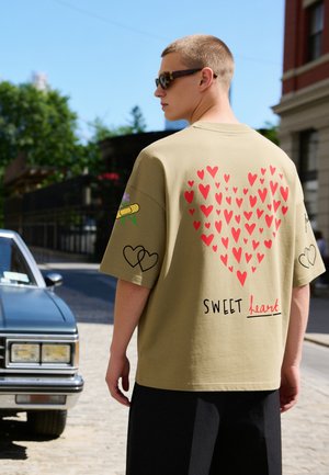 Junger Mann trägt Sonnenbrille, schaut weg, beiges T-Shirt mit roten Herzmustern und dem Text "SWEET heart" auf dem Rücken, steht neben einem Oldtimer im Freien.