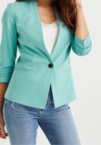 Blazer de color verde claro con cierre de un solo botón, mangas tres cuartos y corte entallado, combinado con una blusa de encaje blanca y jeans.