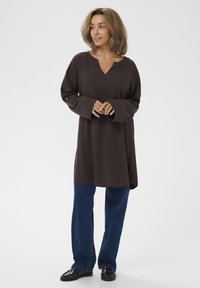 Tunic marrone scuro a maniche lunghe con scollo a V, realizzata in tessuto morbido, abbinata a jeans blu a gamba dritta e mocassini neri.