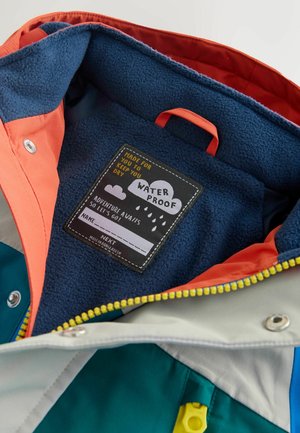 Buntes Innenfutter einer wasserdichten Jacke mit blauem Fleece, orangefarbenem und grauem Stoff sowie einem eingenähten Etikett mit Pflegehinweisen.