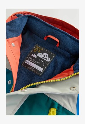 Värvikas veekindla jaku sisemus, kus on sinine fliisvooder, oranž ja hall kangas ning õmmeldud hooldusjuhistega etikett.