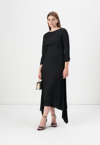 A model wears a black, textured dress with three-quarter sleeves and a flowing hem, holding a small gold handbag and wearing strappy heels.