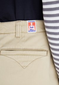 Pantalons Wrangler beiges avec un point de couture contrasté marron, dotés d'une poche arrière et d'une étiquette logo blanche et bleue.