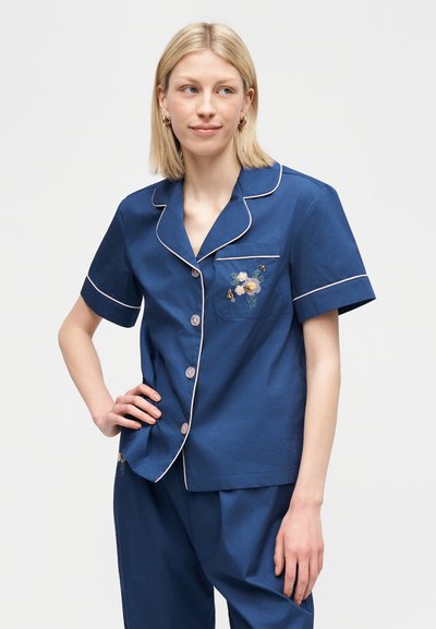 Γυναίκα που φορά σετ πυτζάμες σε σκούρο μπλε με κοντά μανίκια, λευκές ραφές και κεντήματα με λουλούδια στην τσέπη στο στήθος, στέκεται με το χέρι στη μέση.