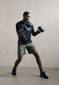 T-shirt noir à manches courtes avec un design géométrique, shorts verts olive avec des accents blancs, gants de boxe noirs et chaussures de sport noires.